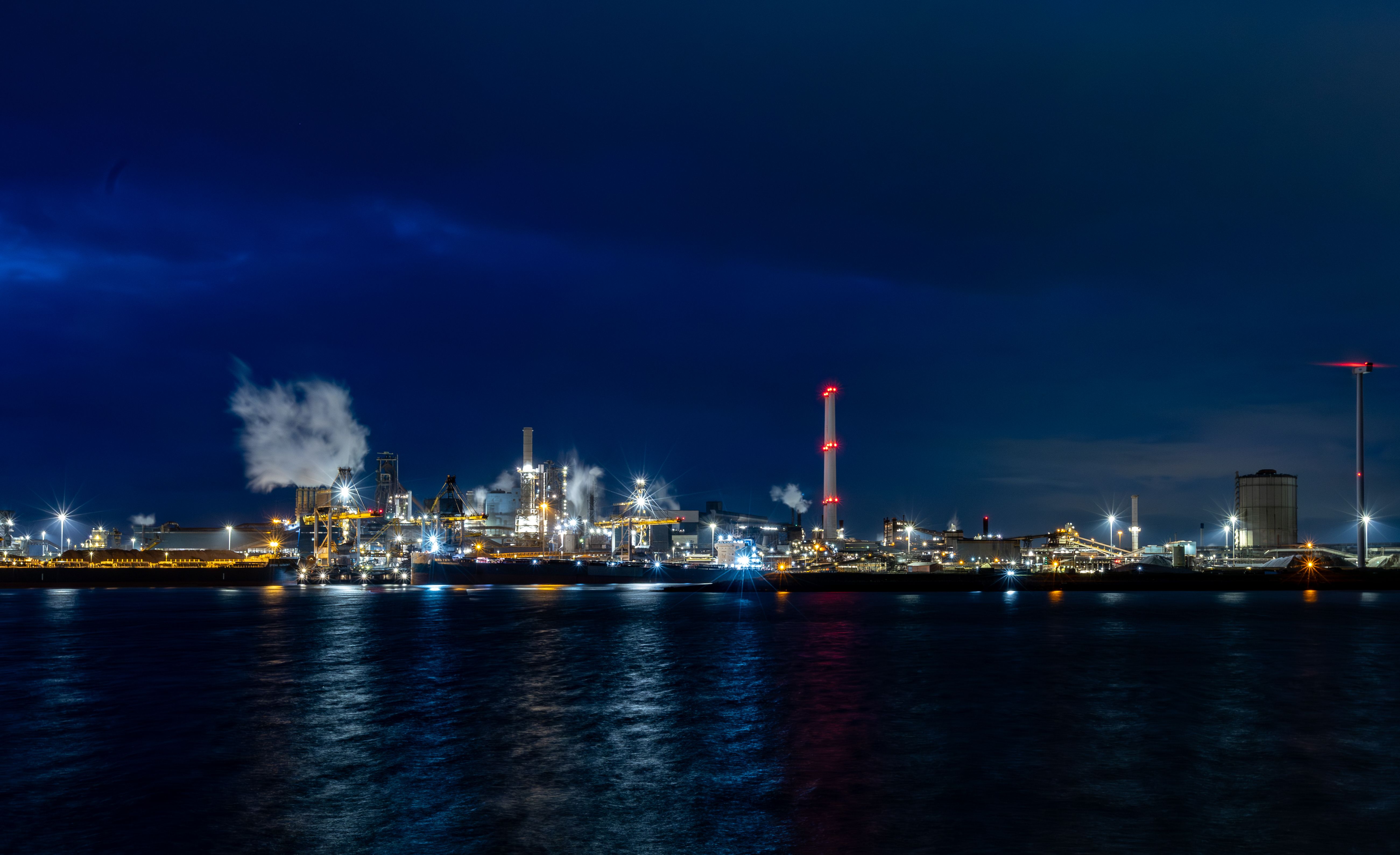 Industrial complex at night along a harbor, with illuminated towers, steam plumes, and light reflections on the water.